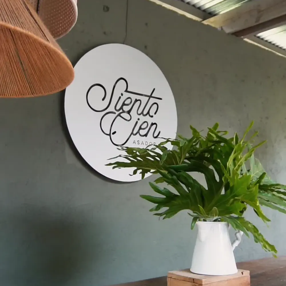 Letrero circular blanco con el nombre Siento Cien Asador escrito a mano, colgado en una pared verde junto a un helecho.