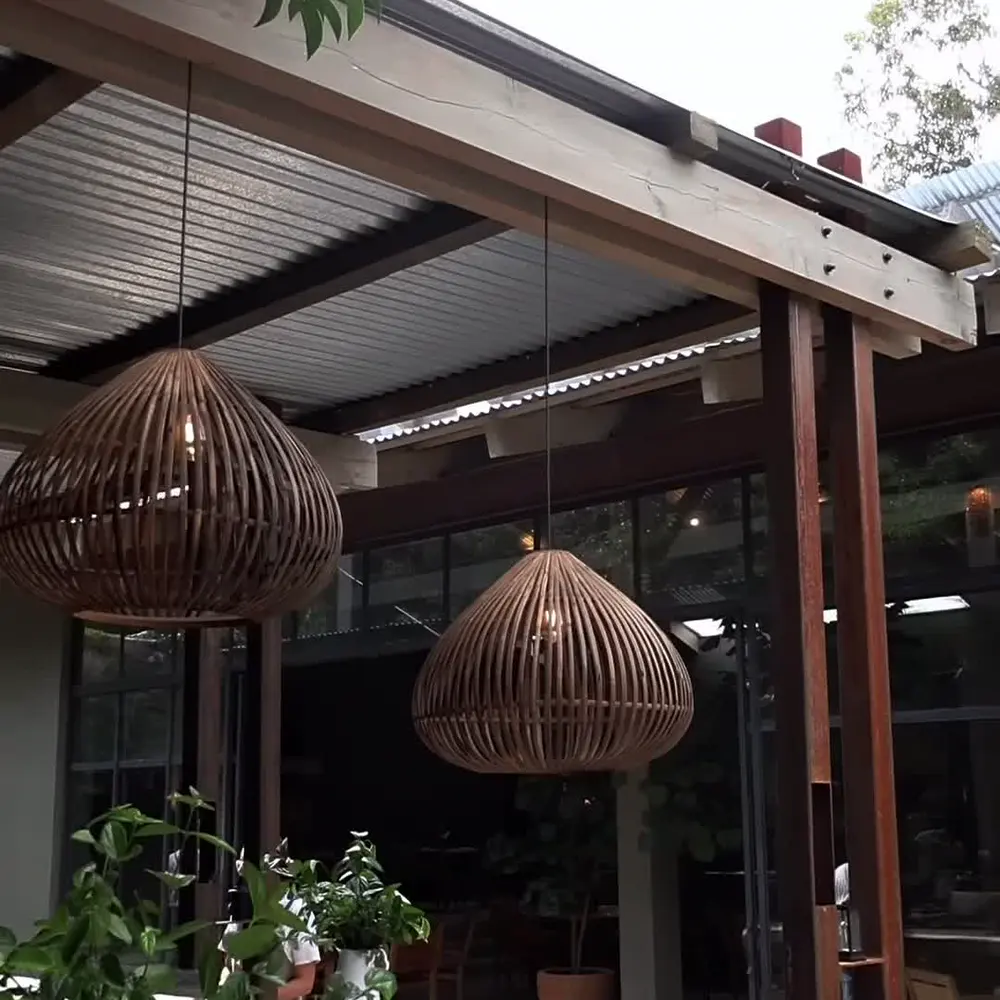 Terraza techada con estructura de lámina y vigas de madera, lámparas de mimbre y plantas al frente.