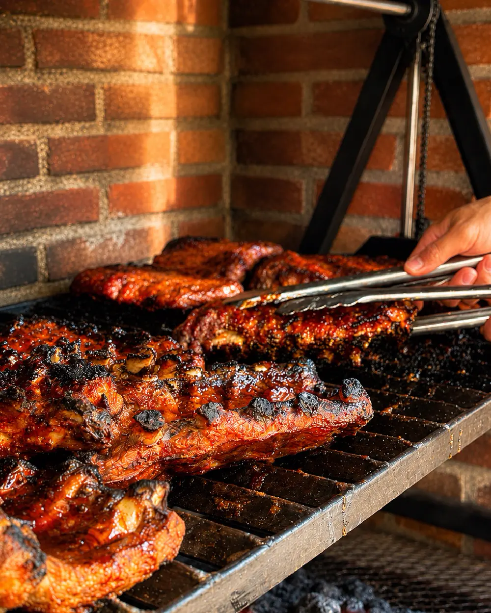 Costillas glaseadas sobre la parrilla de carbón, junto a un muro de ladrillo, sujetas con pinzas.