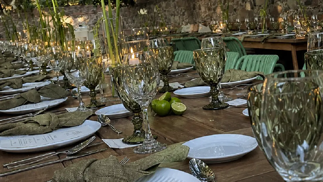 Mesa larga de madera puesta bajo una enredadera, con copas verdes, servilletas de lino y una carta impresa sobre cada plato.